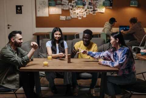A diverse group of young adults laughing while playing a board game together at a table, with language flashcards mixed in with game pieces, suggesting fun learning.